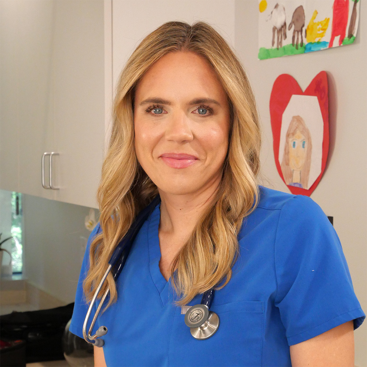 Headshot of Dr. Annie Andrews wearing blue scrubs with children's drawings on the wall behind her.